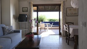 A living room with a sofa, a coffee table with a vase, a television on a stand, and a dining area with a table and chairs. - Aria di Vacanza - Corallina (Loiri Porto San Paolo)