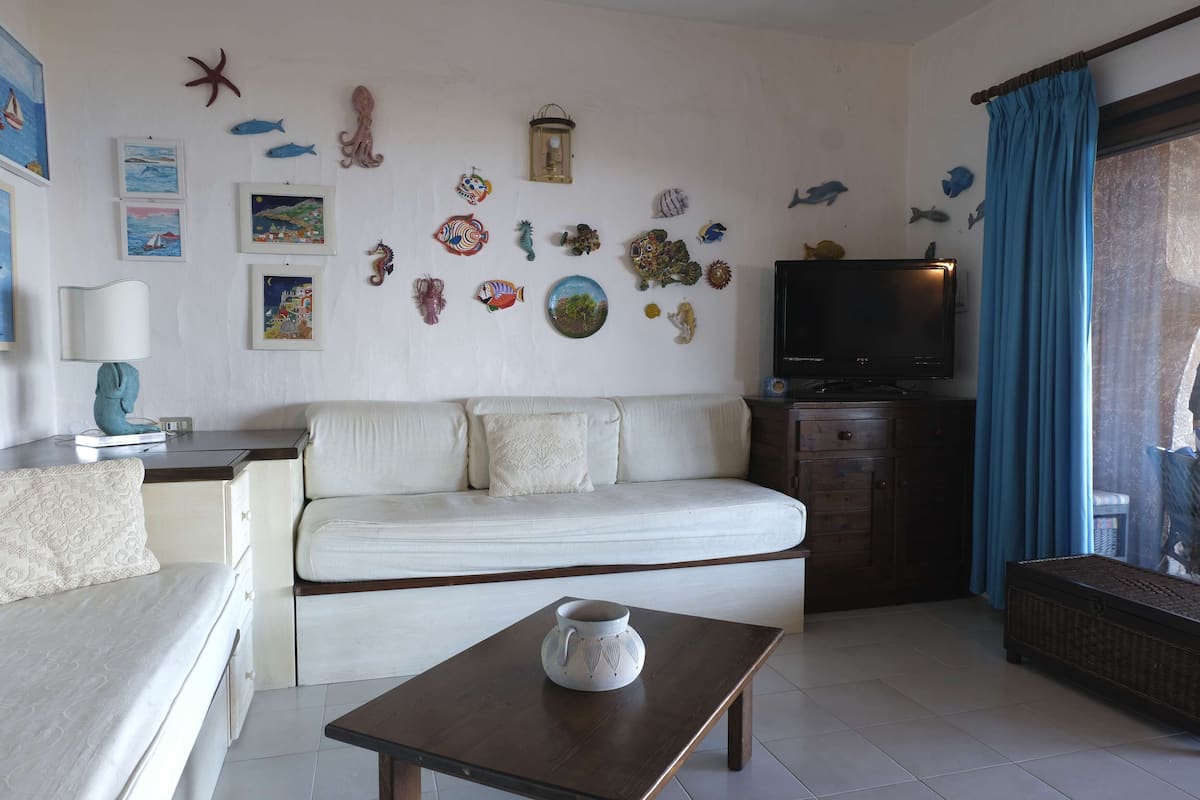 Ein Wohnzimmer mit einem weißen Sofa, einem kleinen Tisch mit einer Dekoration, einem Fernseher und Wandverzierungen, darunter Fische und ein Laternenbild.