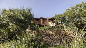 A rustic stone wall and a traditional building with a tiled roof, surrounded by dense greenery. - Aria di Vacanza - Corallina (Loiri Porto San Paolo)