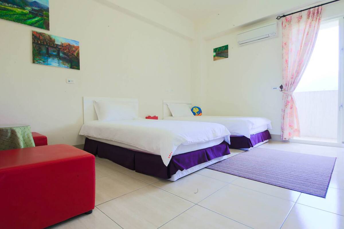 A hotel room with two beds, a red chair, a small table, and a window with curtains.