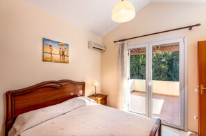 A bedroom with a wooden bed, a nightstand with a lamp, a painting on the wall, and a sliding glass door leading to an outdoor balcony.