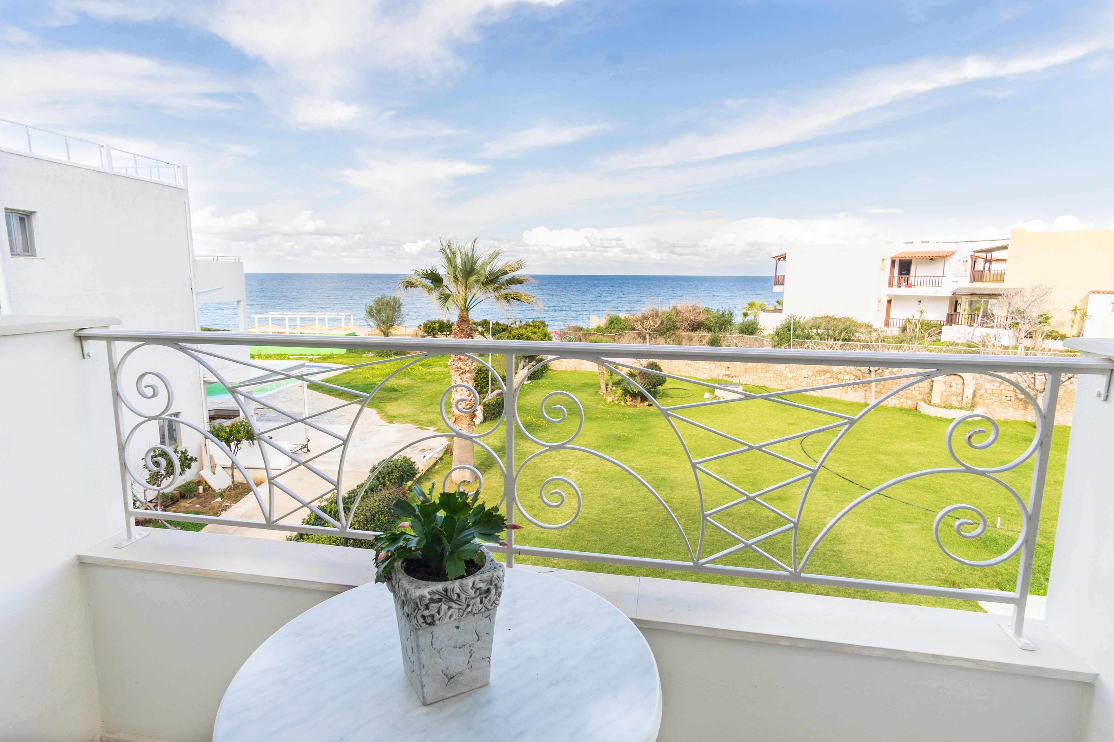 A balcony with a white table and potted plant, overlooking a well-maintained lawn and a view of the sea.