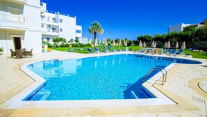 A swimming pool with lounge chairs and umbrellas, surrounded by a well-maintained lawn and a building with balconies. - Pela Sofia (Hersonissos)