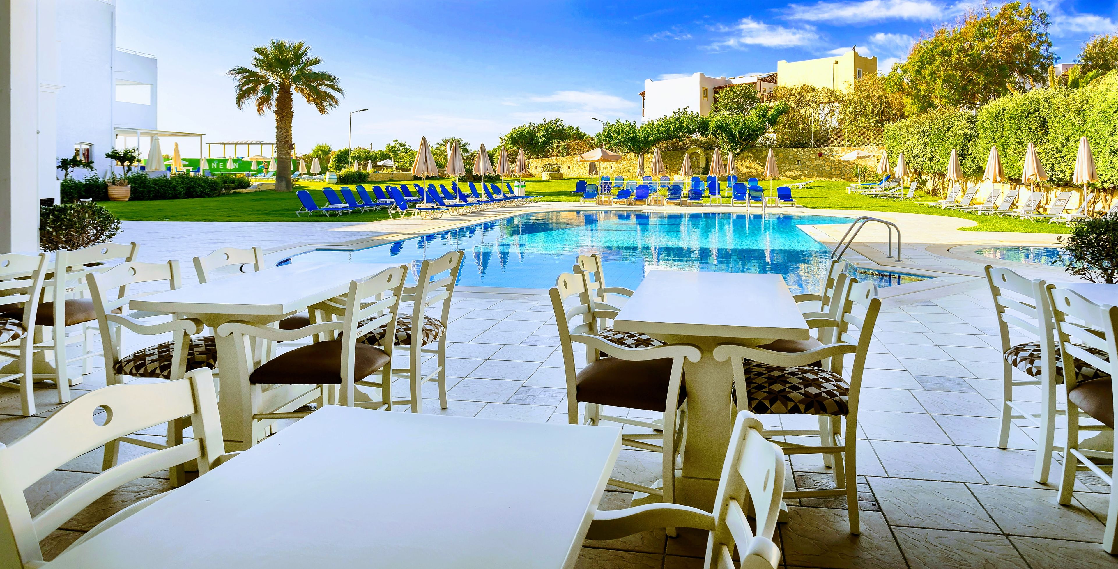 A poolside area with white tables and chairs, a clear blue pool, and a green lawn with lounge chairs and umbrellas.