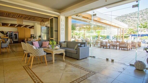 A modern hotel lobby with a sofa, chairs, a coffee table, and a glass wall offering a view of the outdoor seating area. - Pela Sofia (Hersonissos)