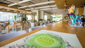 A modern restaurant with wooden tables and chairs, a long counter, and hanging pendant lights. - Pela Sofia (Hersonissos)
