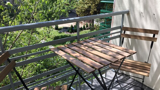 Une table et des chaises en bois sur un balcon doté d’une rambarde en métal, donnant sur un jardin avec des arbres et un bâtiment en arrière-plan.