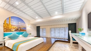 Quarto de hotel com uma cama grande, uma escrivaninha, uma televisão e vista para uma cidade costeira através de uma janela ampla.