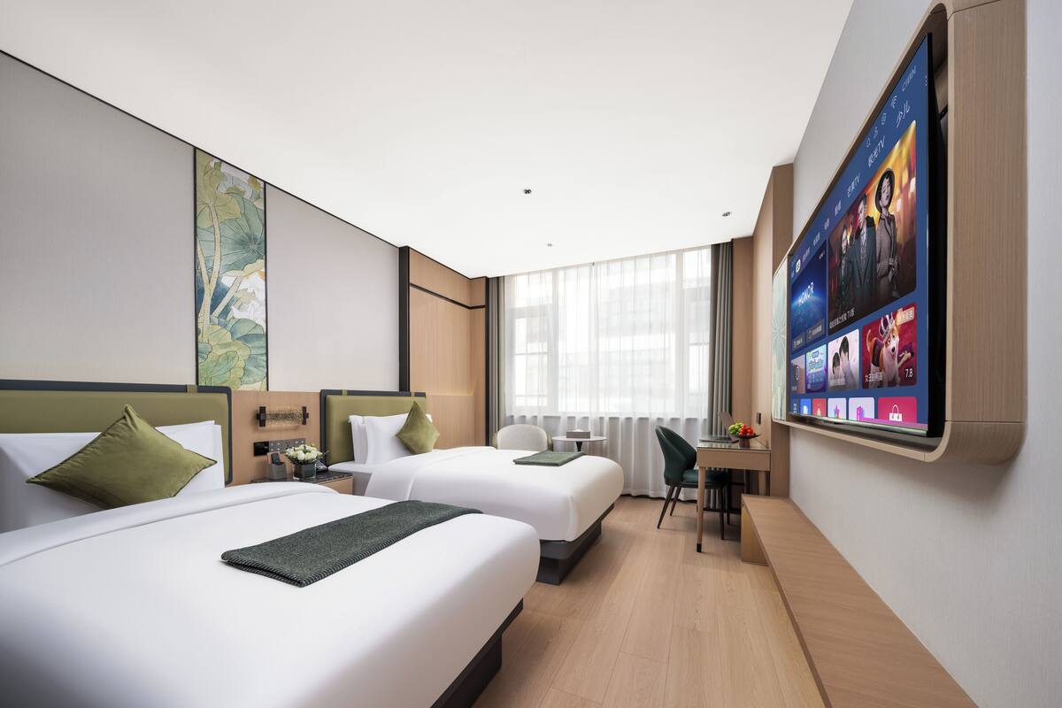 A hotel room with two beds, a desk, a chair, a TV mounted on the wall, and a large window.