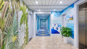 Corredor de hotel com um mural tropical, carpete azul e um vaso de planta.