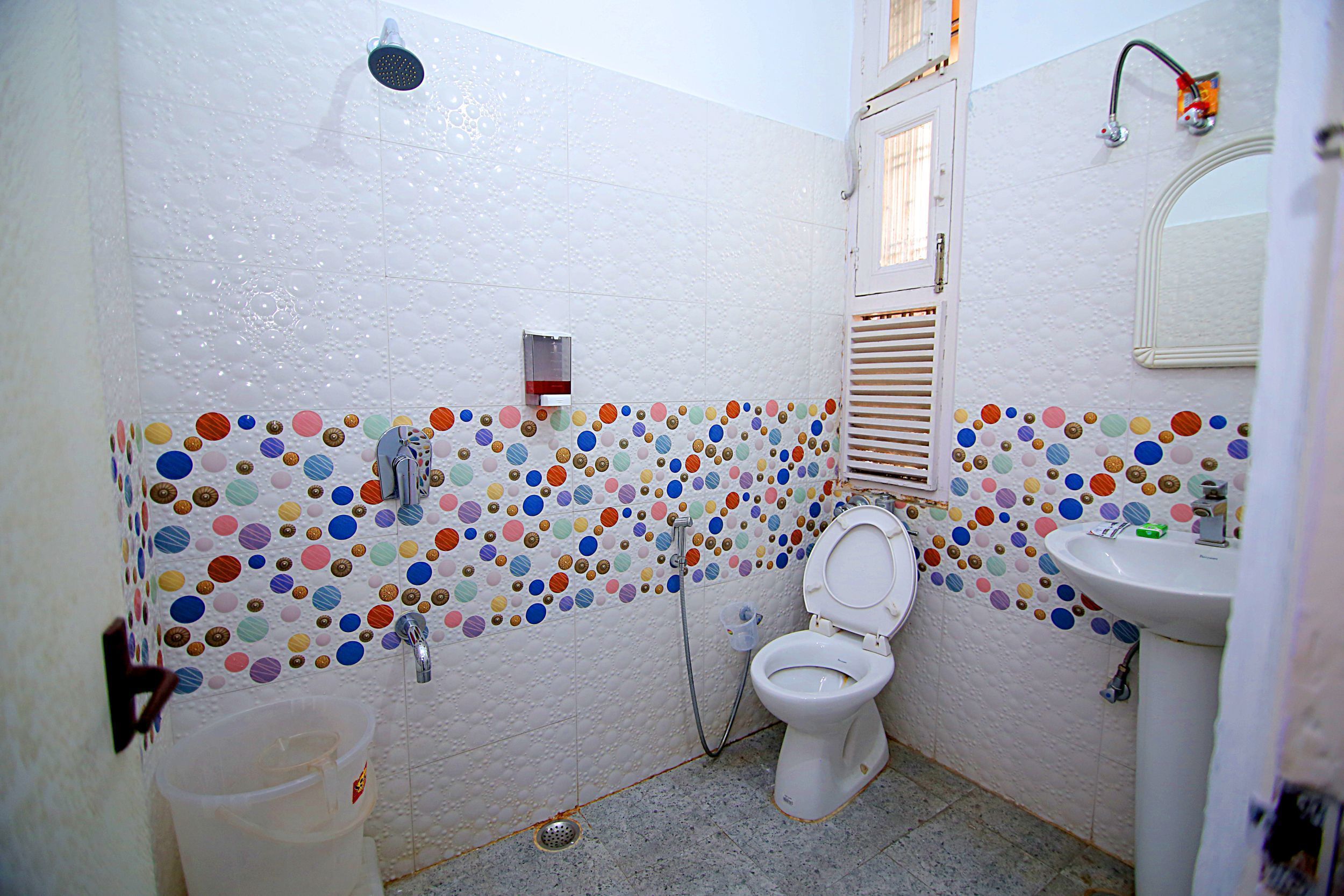 Une salle de bain comprenant des toilettes, un lavabo, une douche et un mur recouvert de carreaux circulaires aux couleurs vives.