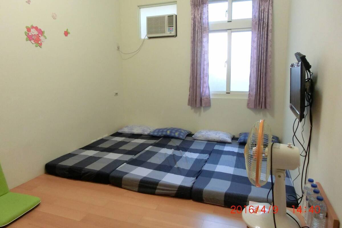 A small room with a bed, a window with curtains, a wall-mounted air conditioner, a television, and a standing fan.