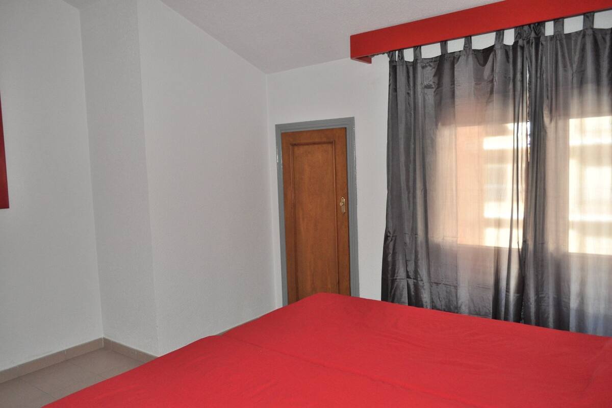 A room with a red bedspread, a wooden door, and a window with grey curtains.