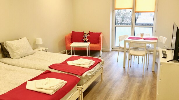 A hotel room with two beds, a small table, a chair, a TV, and a window with blinds. - VisitLublin Apartments Premium Wschodnia (Lublin)
