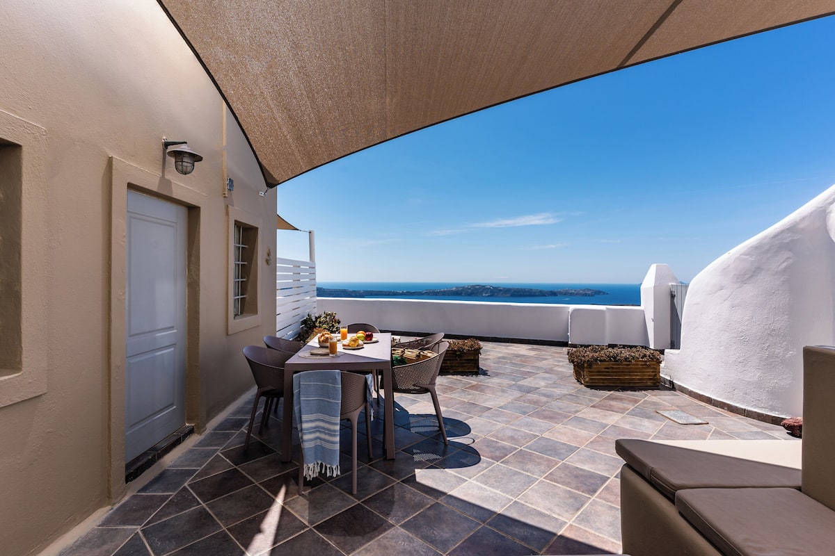 Una terraza con una mesa para dos, una silla y un sofá. Se ve el mar y un edificio al fondo.