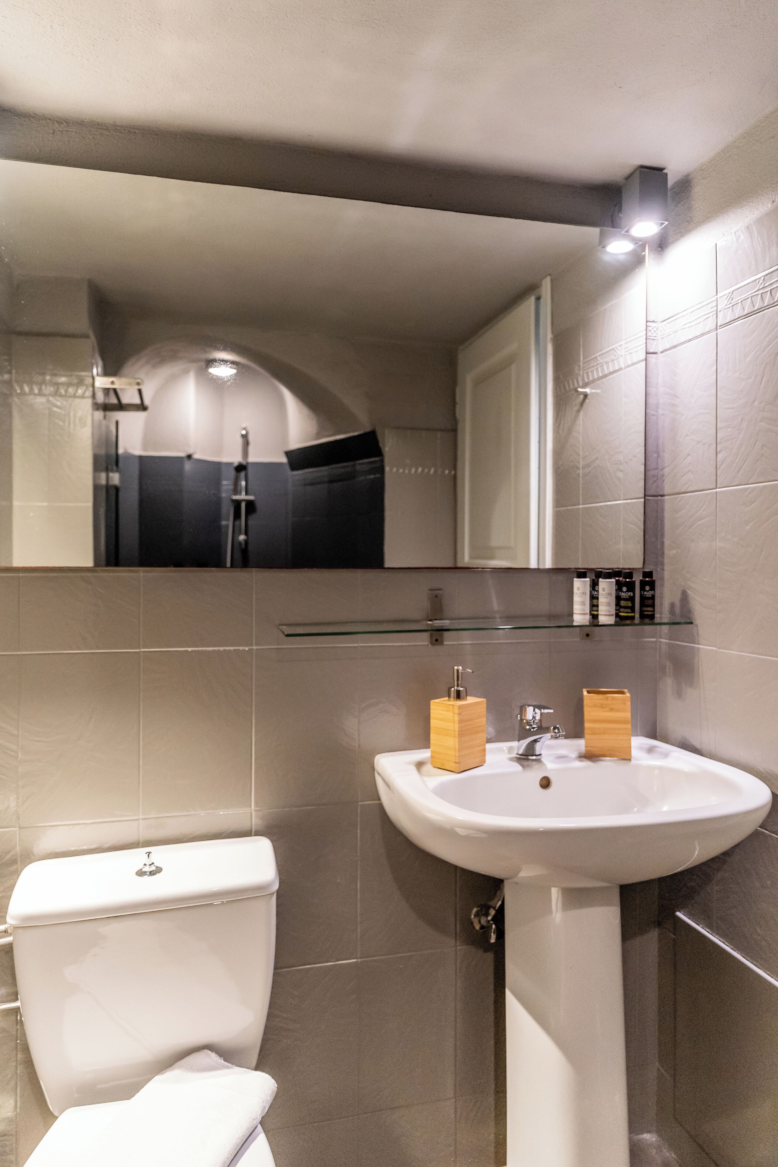 A bathroom with a white sink, wooden soap dispensers, a toilet, and a glass shelf with toiletries.