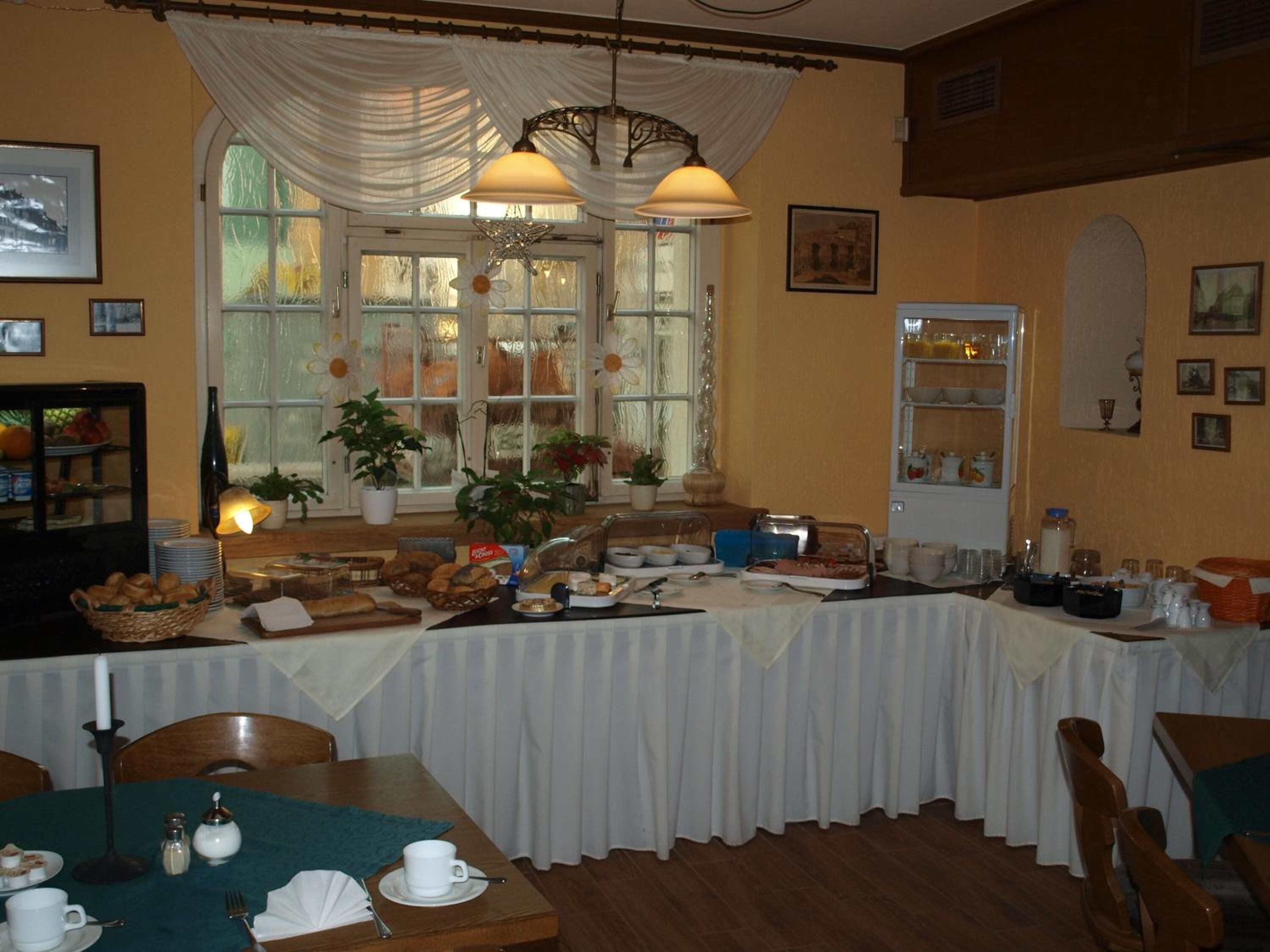A dining area with a buffet setup, including bread, pastries, and various dishes. There are dining tables, chairs, and a large window with curtains.