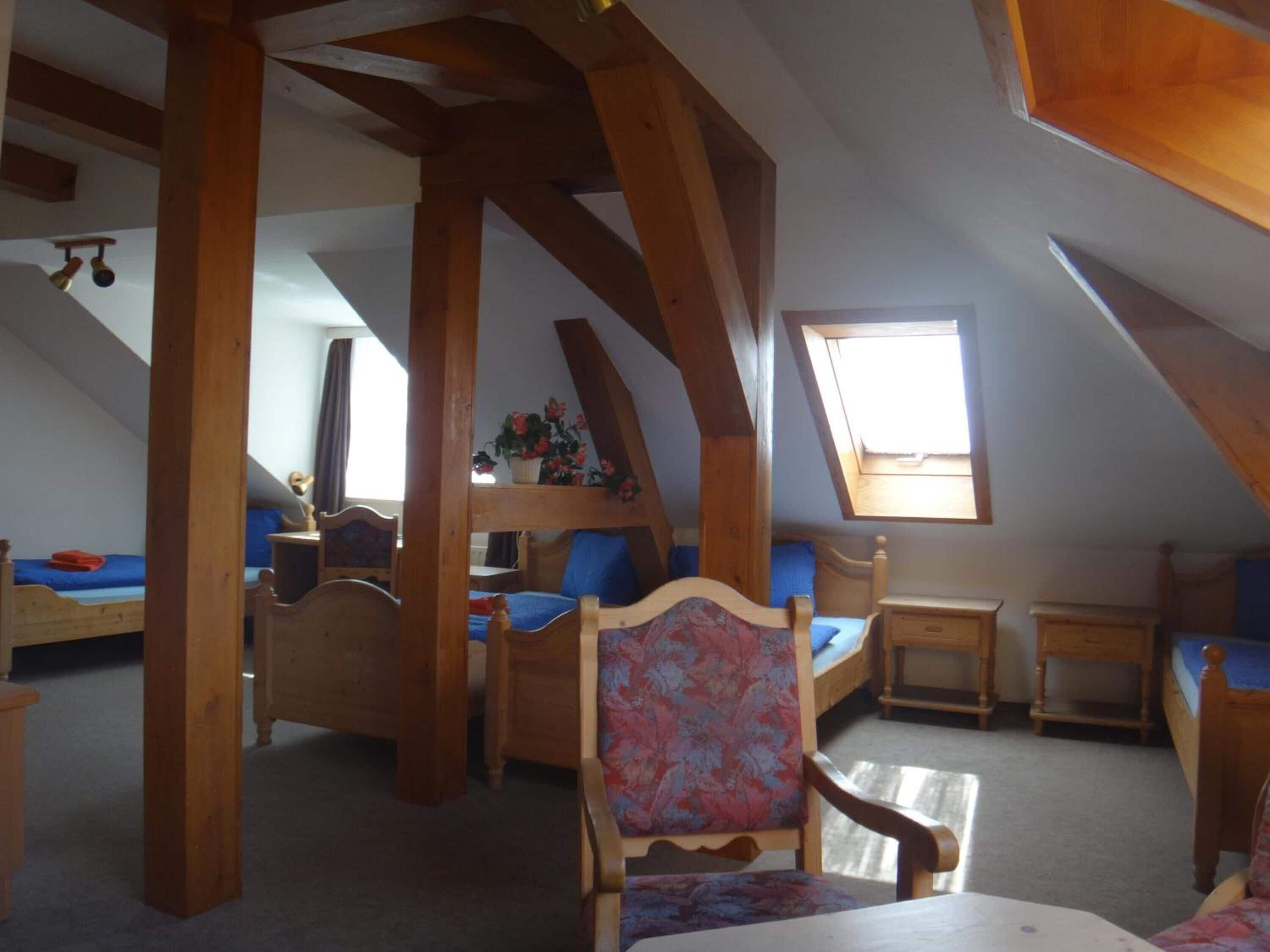Um quarto de sótão aconchegante com vigas de madeira aparentes, teto inclinado e um domo. Há duas camas, uma cadeira e uma mesa pequena.