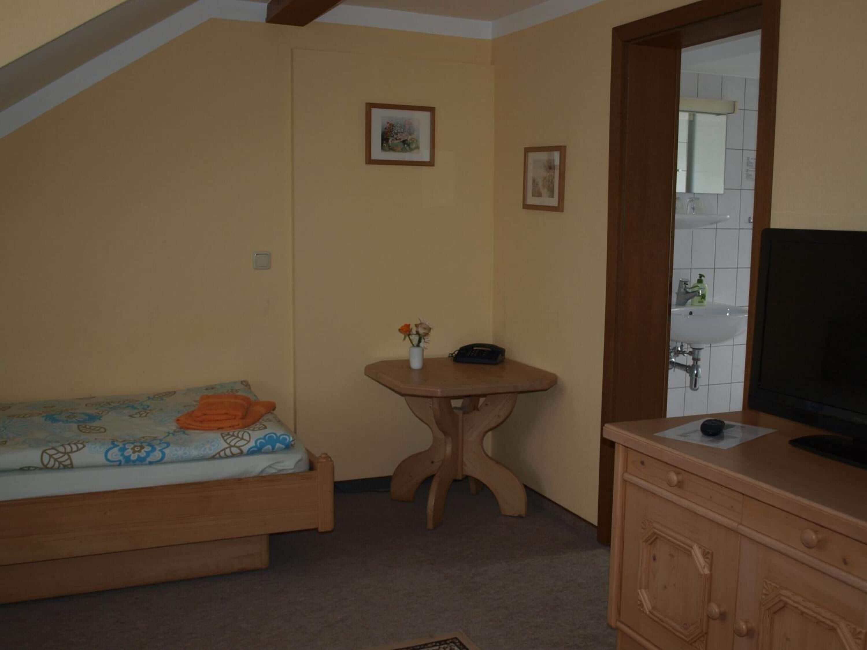 A bedroom with a bed, a small wooden table with a vase and a phone, a wooden dresser, and a bathroom visible through an open door.