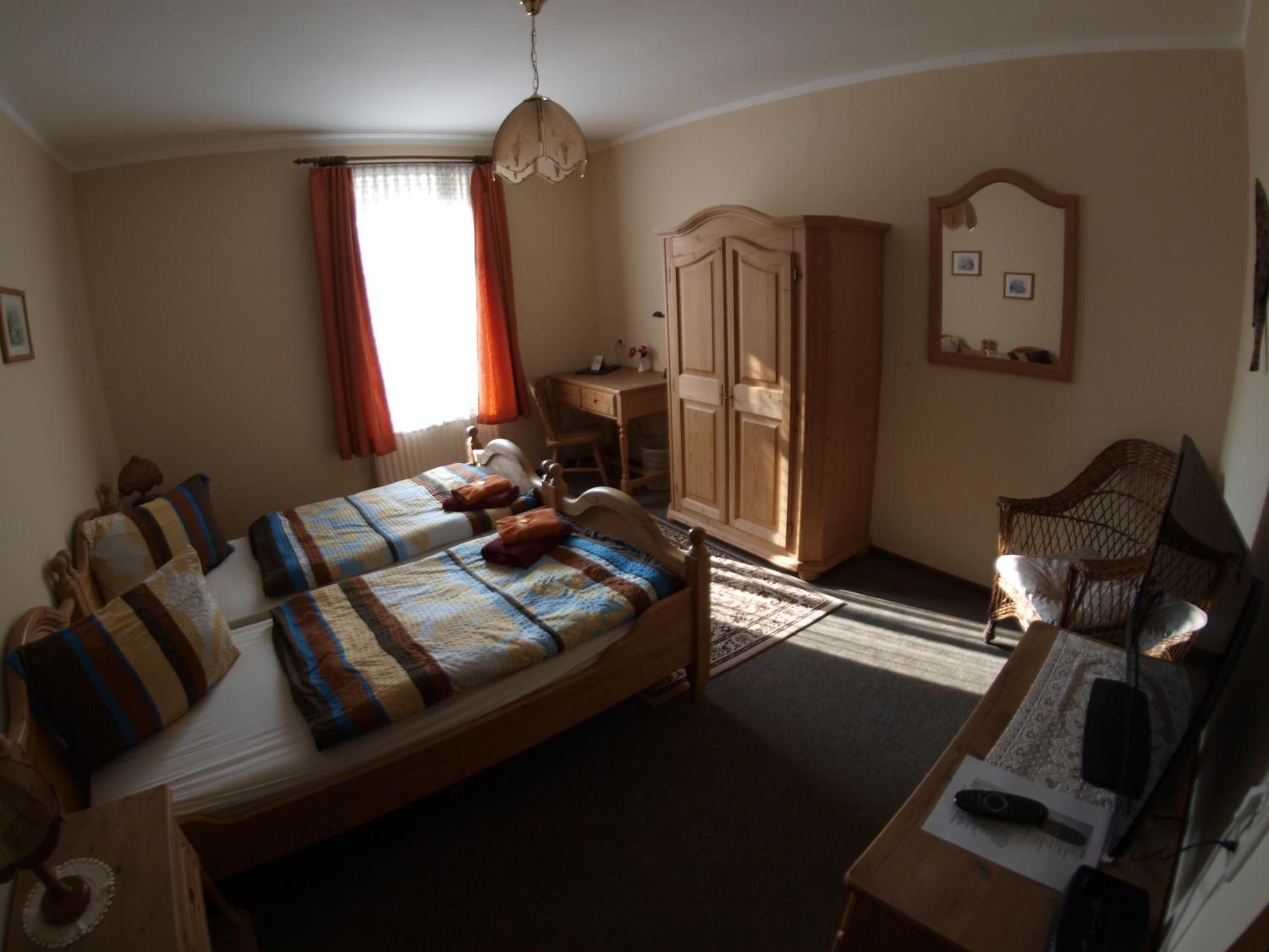 A bedroom with two single beds, a wooden wardrobe, a desk with a chair, a wicker chair, and a mirror on the wall.