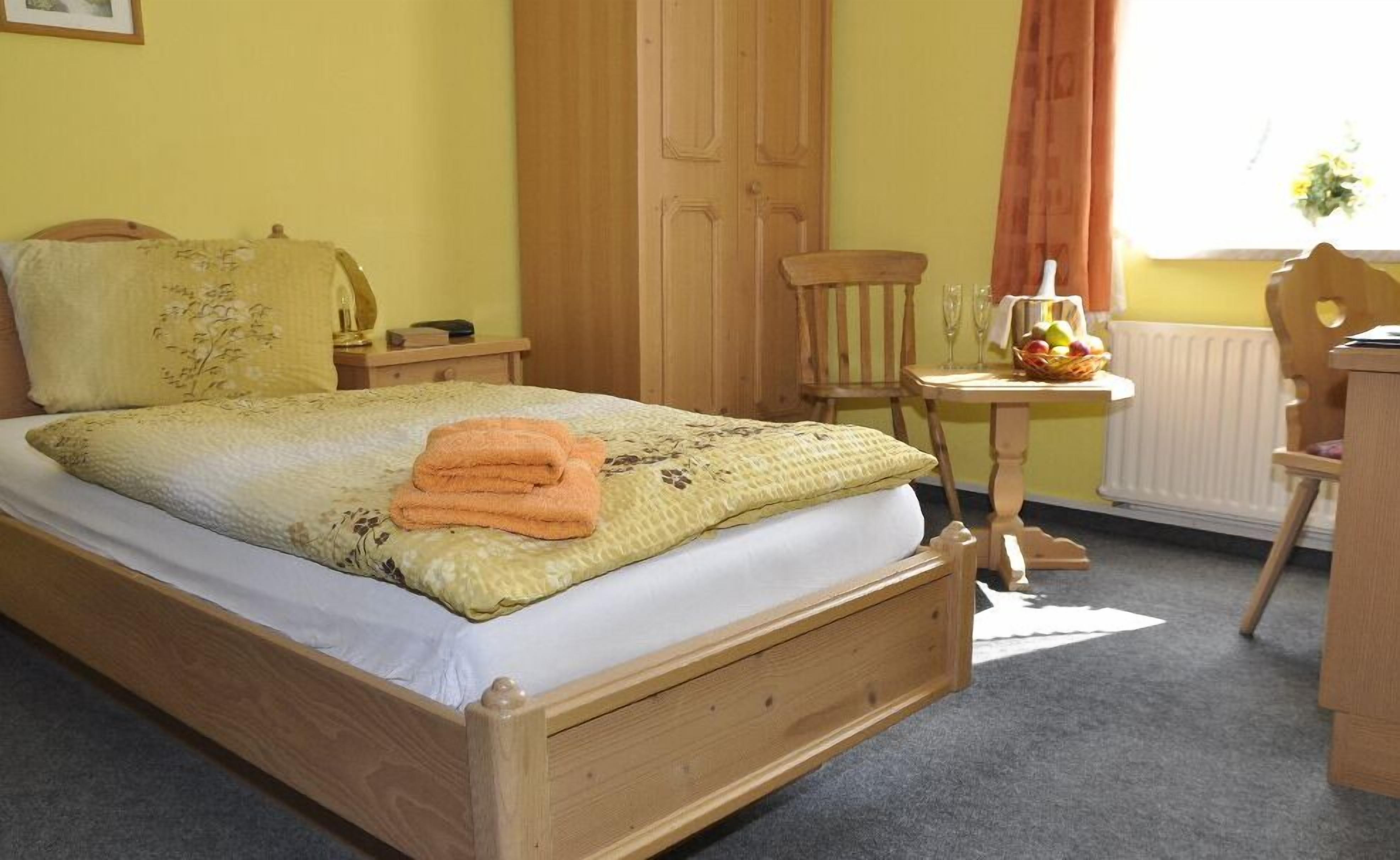 Una cama de madera con colchón blanco y colcha estampada de flores amarillas. Hay una mesita de noche de madera con una lámpara y un teléfono. Se ve una silla de madera y una pequeña mesa de madera con una cesta de fruta.