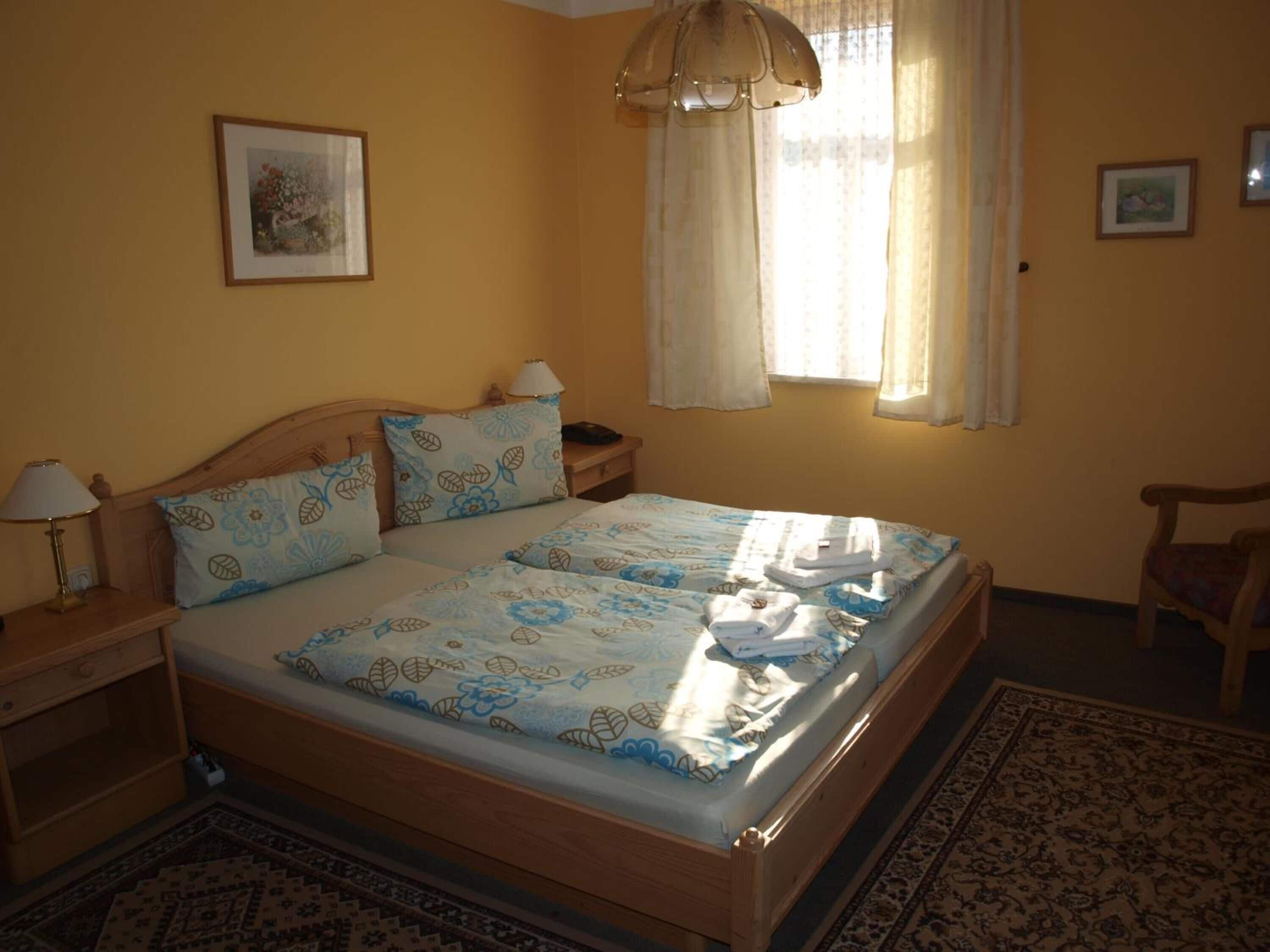 Un lit bien fait, avec une literie à motifs floraux, une tête de lit en bois, une table de chevet avec une lampe, une chaise et une fenêtre avec des rideaux.