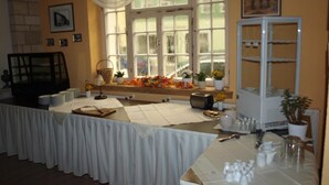 Un comedor con una mesa larga cubierta con mantel blanco, un montaje tipo bufé con platos y cubiertos, y un ventanal con vistas a la vegetación exterior.
