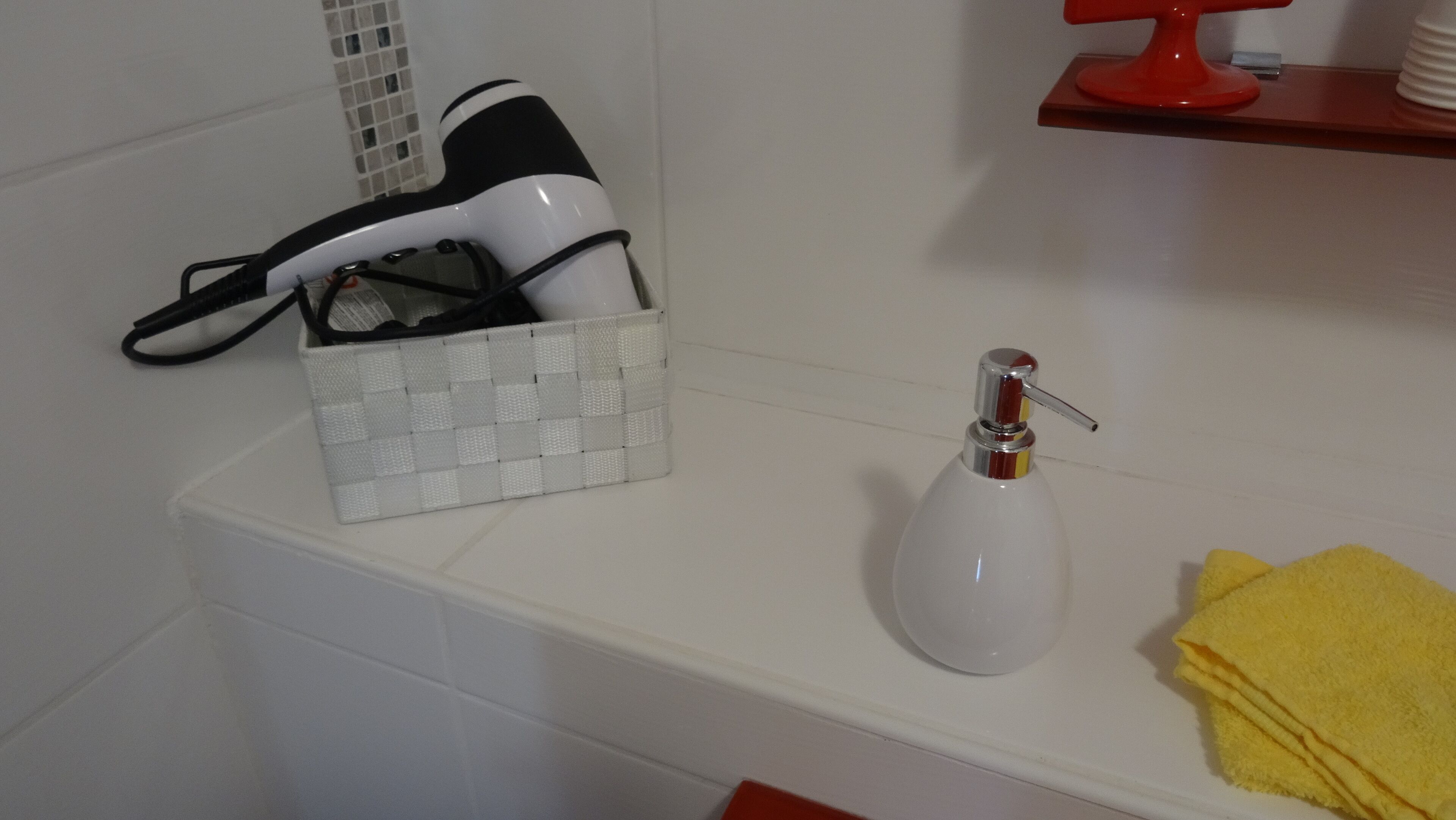 Un baño con un lavamanos blanco, un dispensador de jabón, una toalla amarilla y un zapatero con una cesta blanca que contiene un zapato blanco y negro.