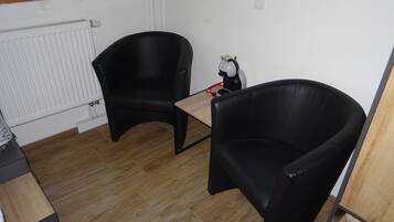 Een kleine kamer met twee zwarte leren stoelen, een houten tafel, een radiator en een plank met een fles en een decoratief object.