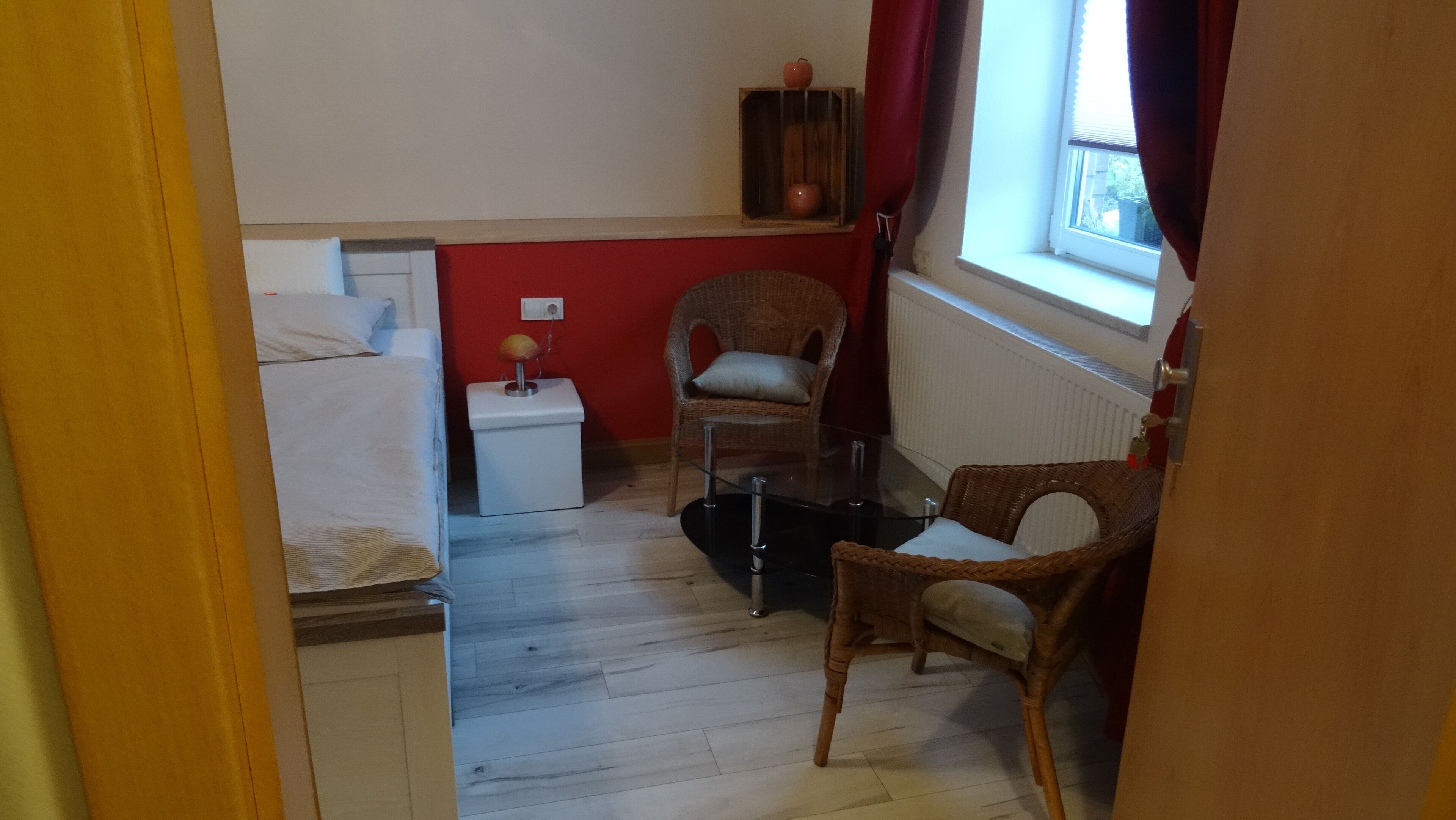 Et lille, kompakt rum med en seng, en stol, et lille bord og et vindue med gardiner.