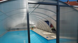 Ein überdachter Pool mit Glaskuppel, Steinböden und Blick auf ein Gebäude mit rotem Dach im Hintergrund.