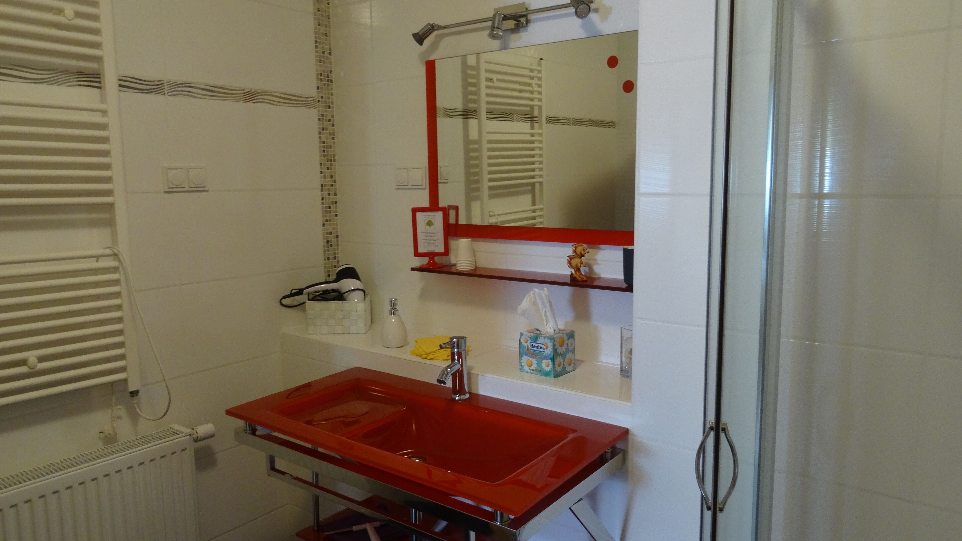 A bathroom with a red sink, a mirror, a towel dispenser, and a small shelf with toiletries.