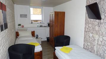 Una habitación pequeña y luminosa con dos camas individuales, un armario de madera, una kitchenette y un televisor montado en la pared.