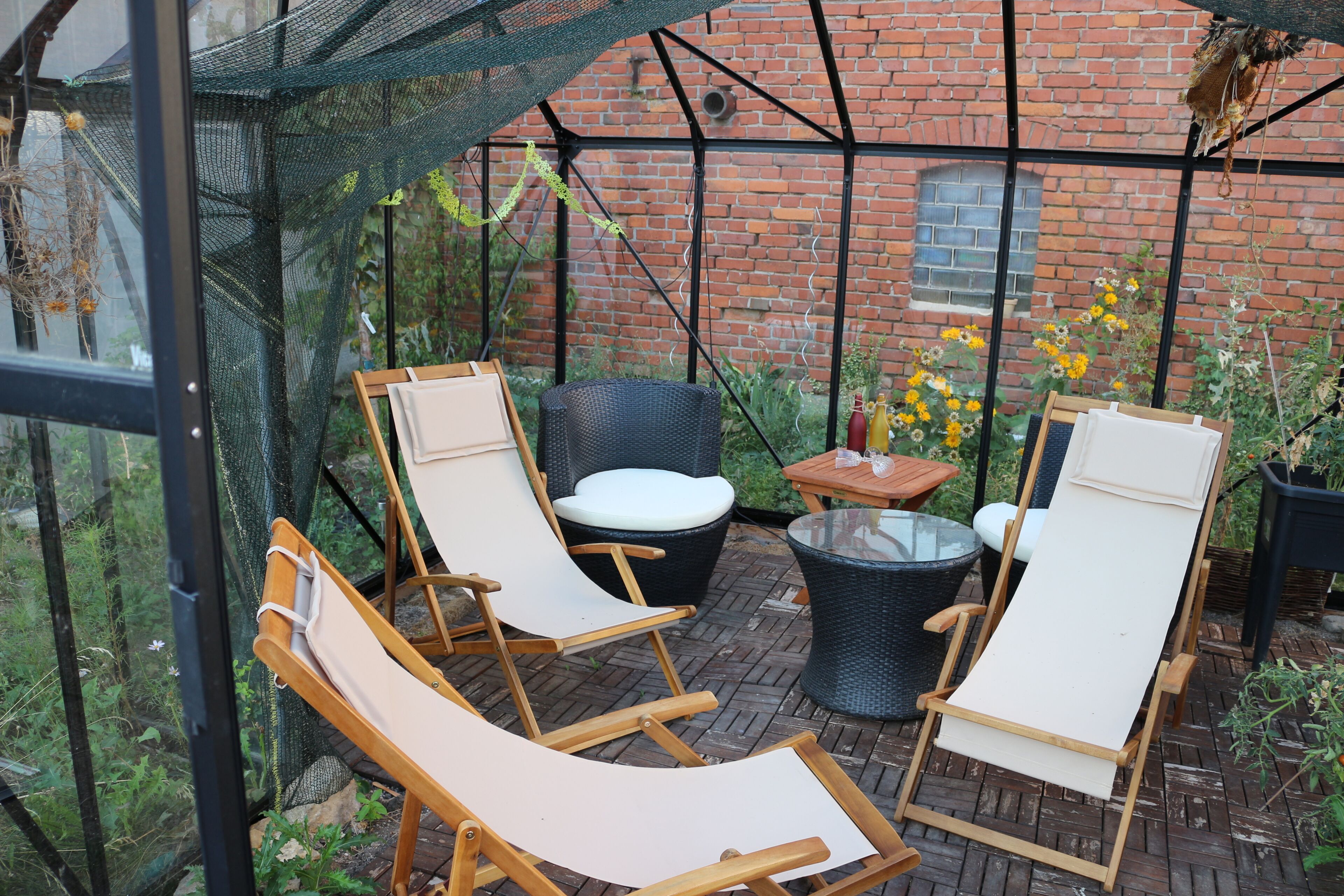 A covered patio area with wooden lounge chairs and a table, surrounded by potted plants and a brick wall.