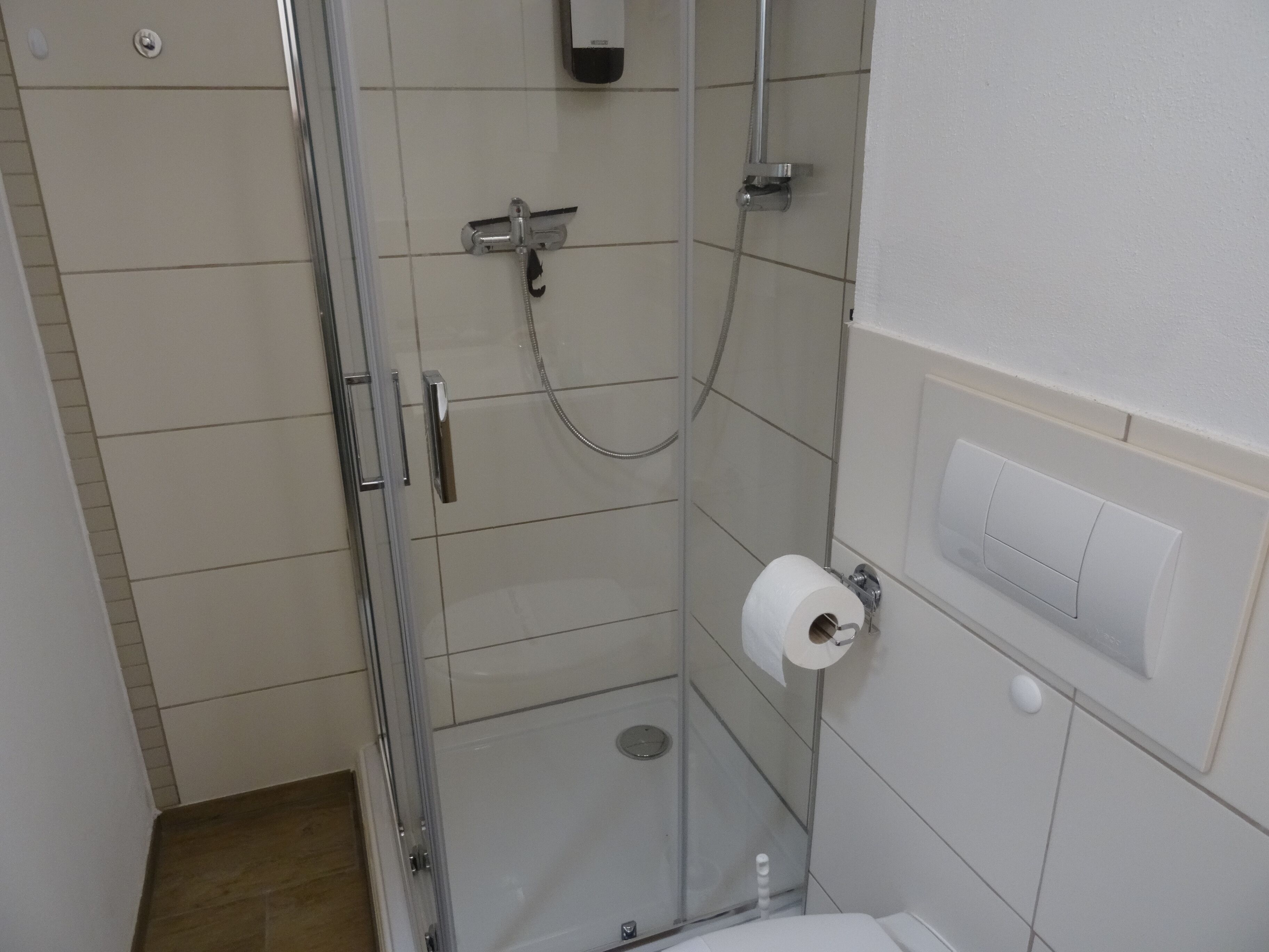 A small, enclosed shower stall with a glass door, a toilet paper holder, and a wall-mounted light switch.