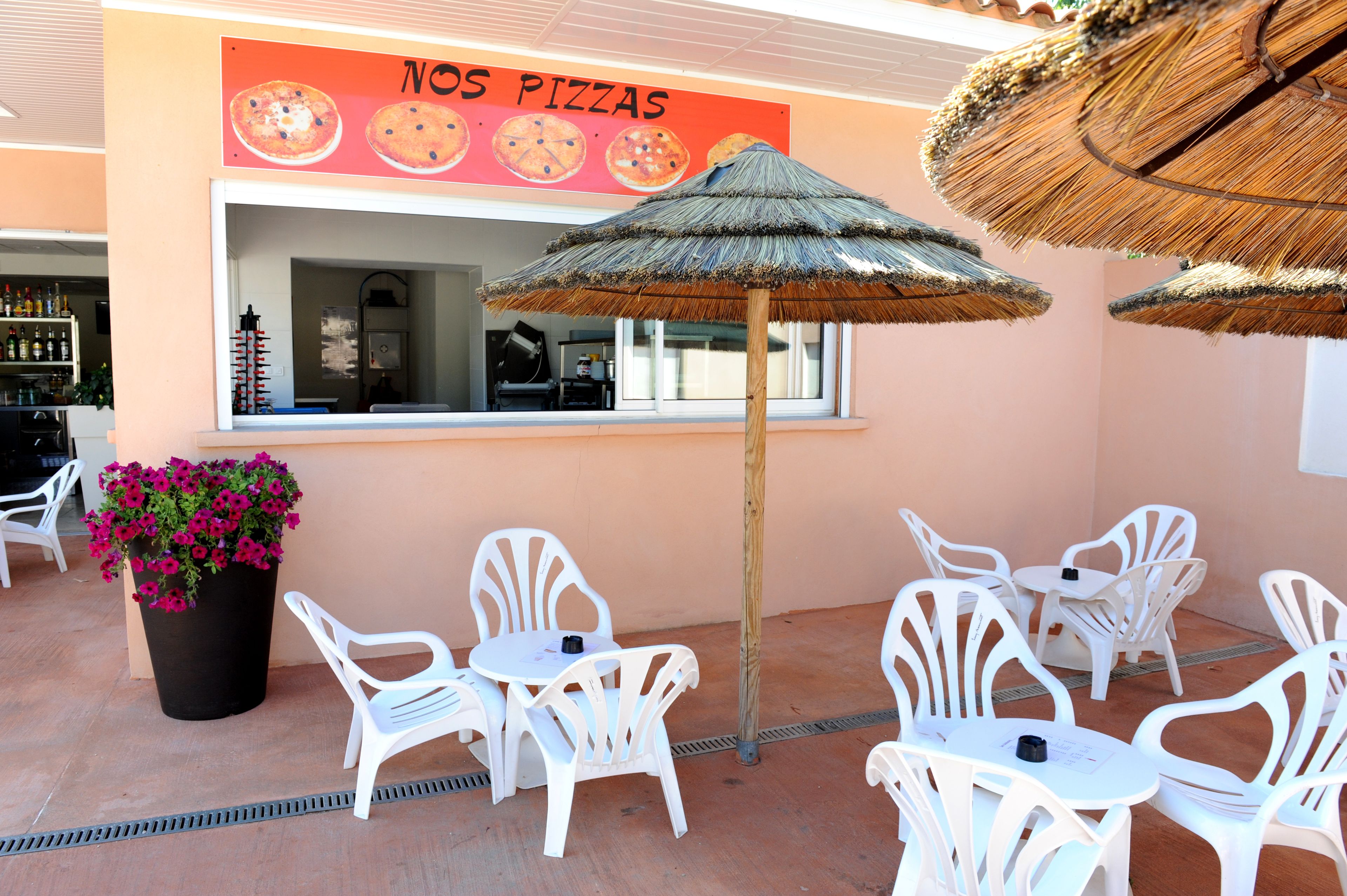 An outdoor seating area with white plastic chairs and tables, a large potted plant with pink flowers, and a thatched umbrella.