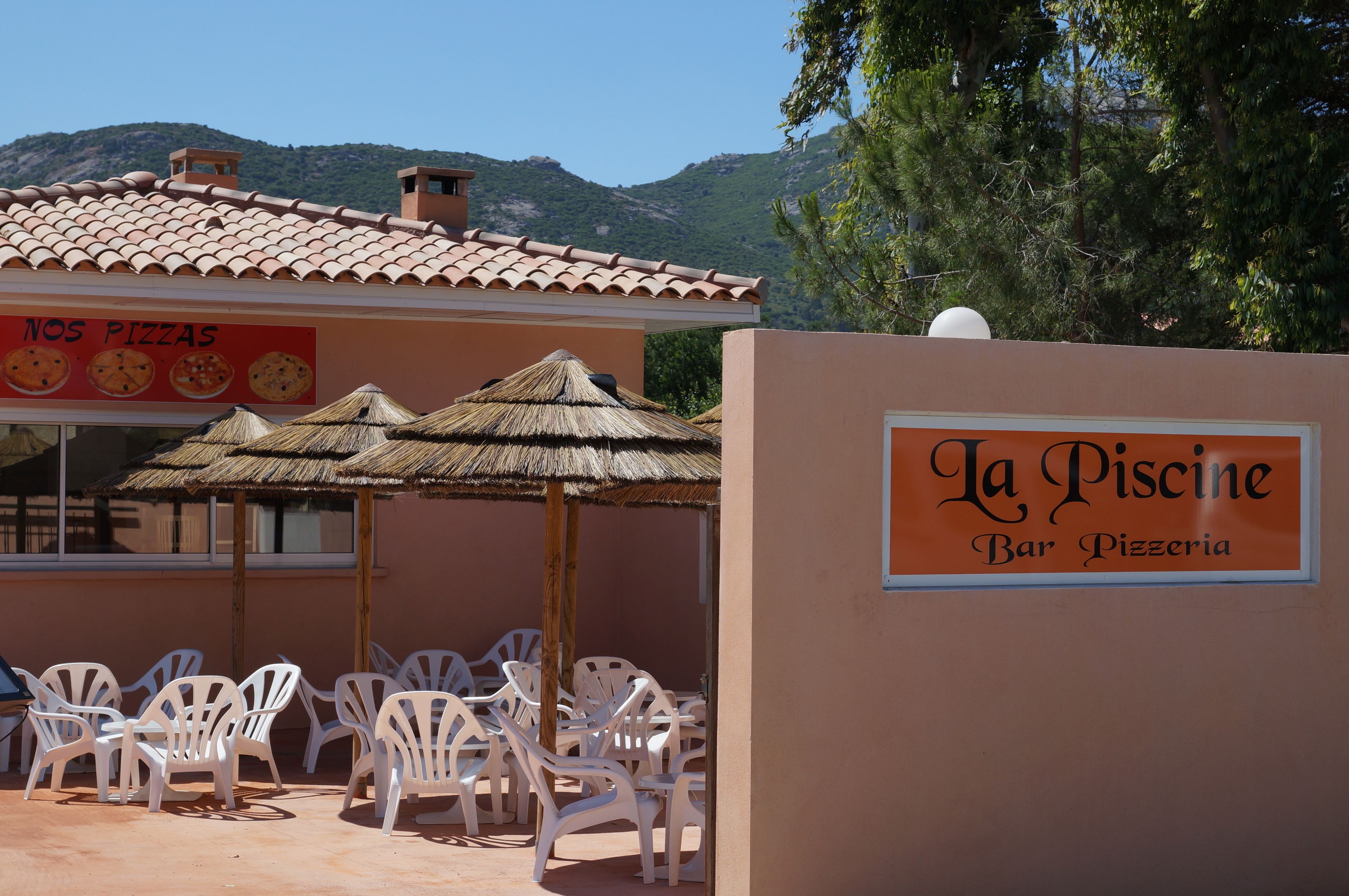 A bar and pizzeria named La Piscine with outdoor seating and thatched umbrellas.