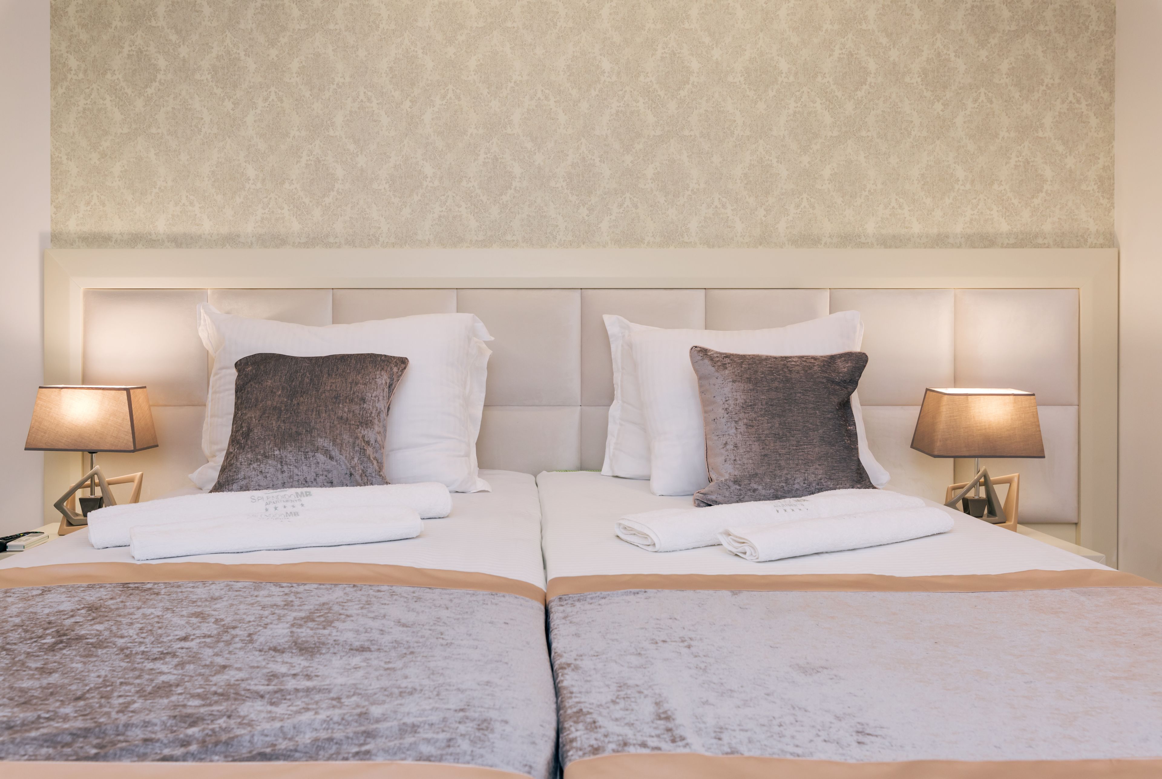 Una cama doble con ropa de cama blanca y marrón, dos lámparas de noche y una pared estampada.