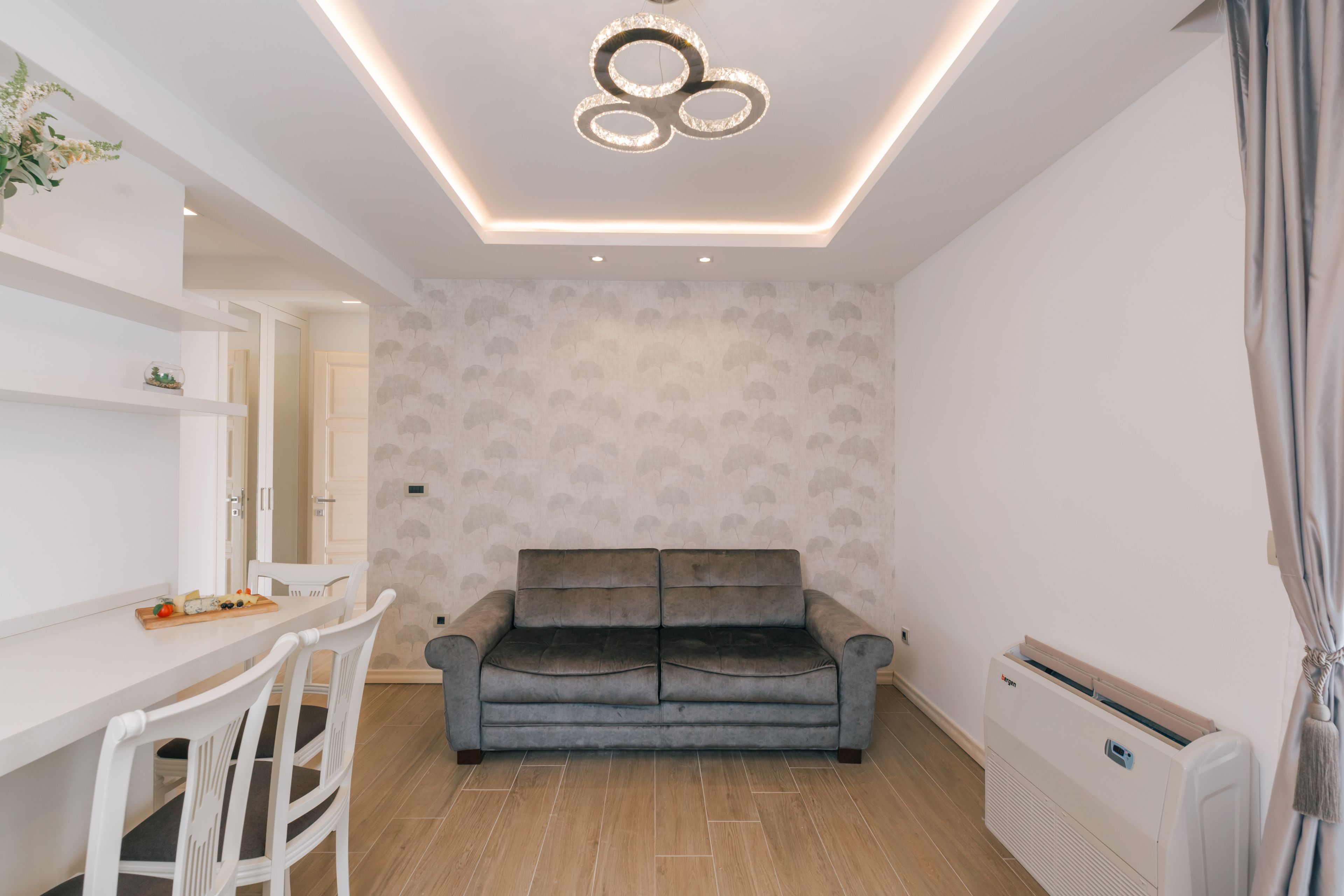 Una sala moderna con un sofá gris, sillas blancas, una mesa de comedor con una bandeja de frutas y un lámpara colgante.