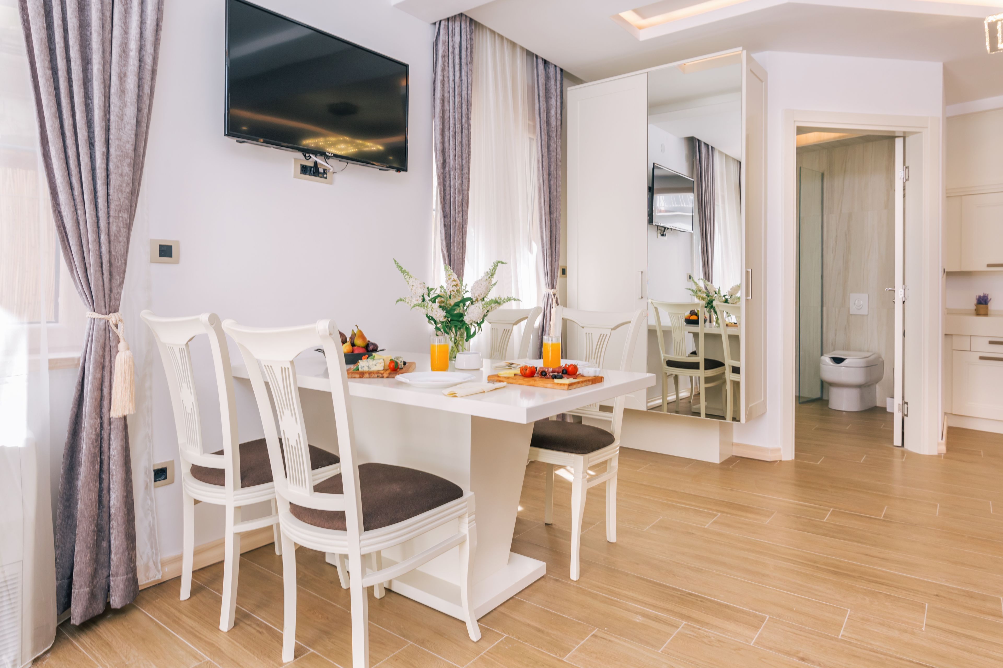 A modern dining area with a white table and chairs, a flat-screen TV mounted on the wall, and a large mirror reflecting the room.