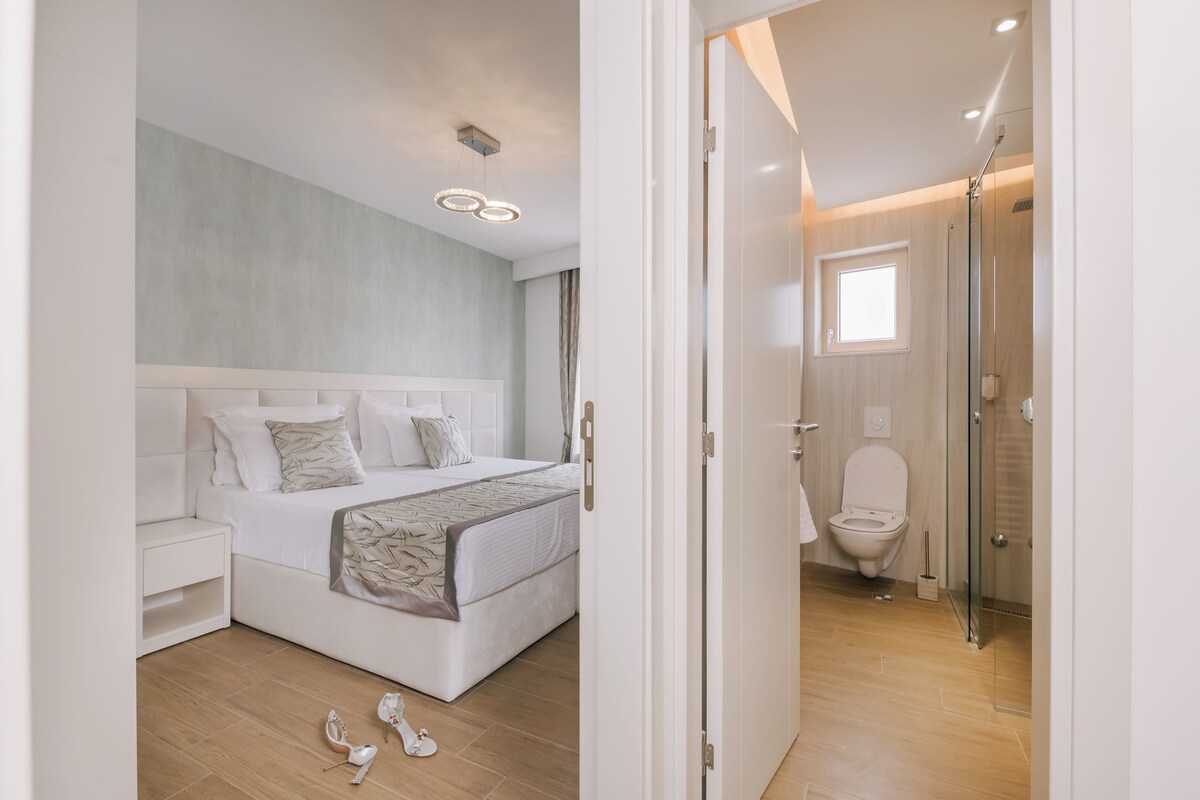 A modern bedroom with a large bed, bedside tables, and a nightstand. There is a bathroom with a toilet, shower, and a window.