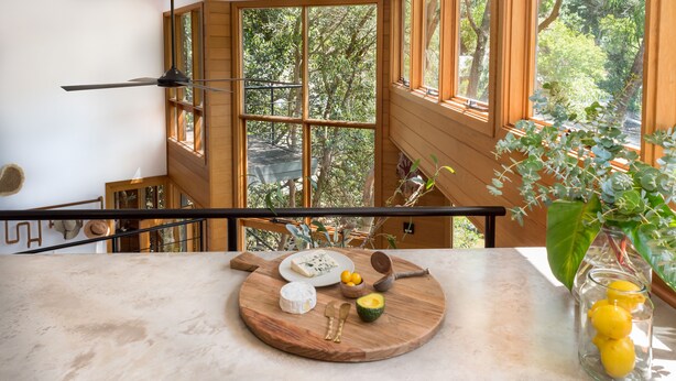 A kitchen counter with a wooden board displaying cheese, a knife, a spoon, and a bowl of fruit.
