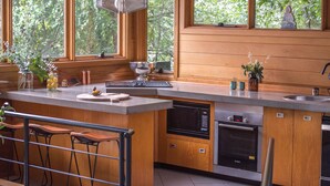 A modern kitchen with wooden cabinets, a large island with bar stools, a built-in oven, and a view of greenery through large windows.