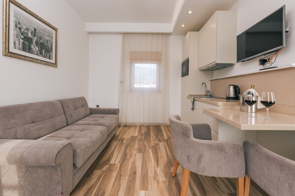 A compact living space with a sofa, two chairs, a kitchen area with a TV, and a window with curtains.