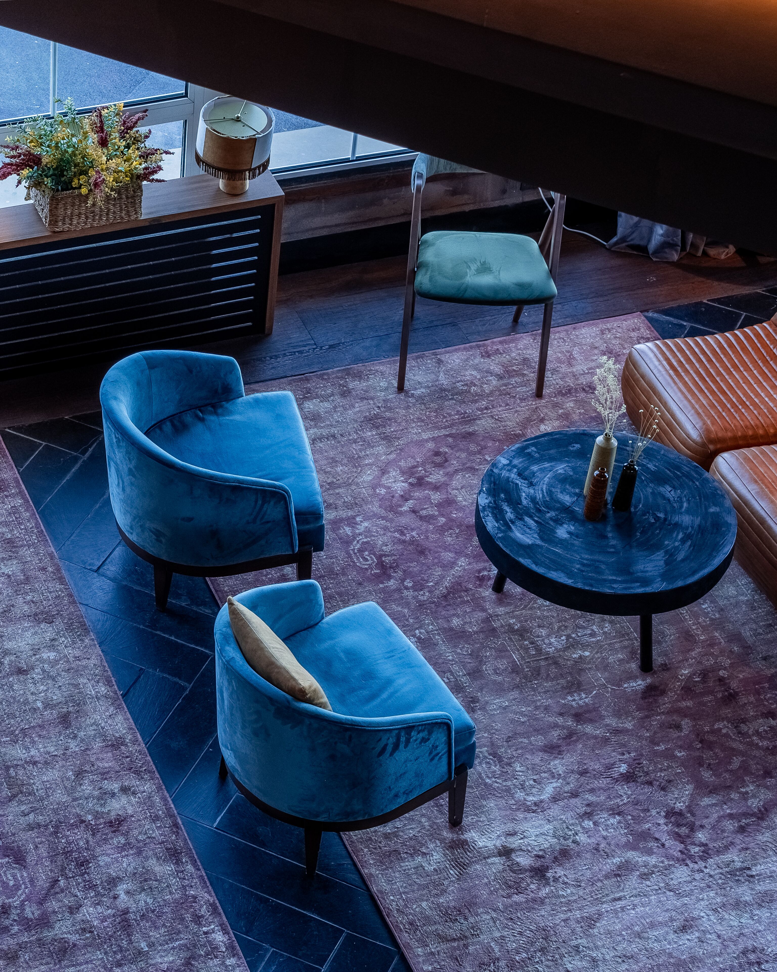 A modern lounge area with blue velvet armchairs, a round coffee table, and a green cushioned chair. A wicker basket with flowers is on a wooden table near a window.