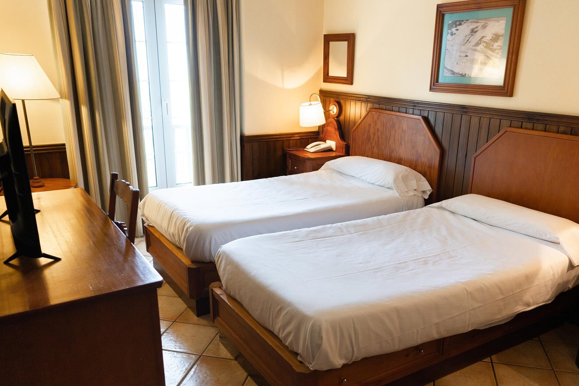 A hotel room with two beds, a desk, a lamp, a telephone, and a framed picture on the wall.