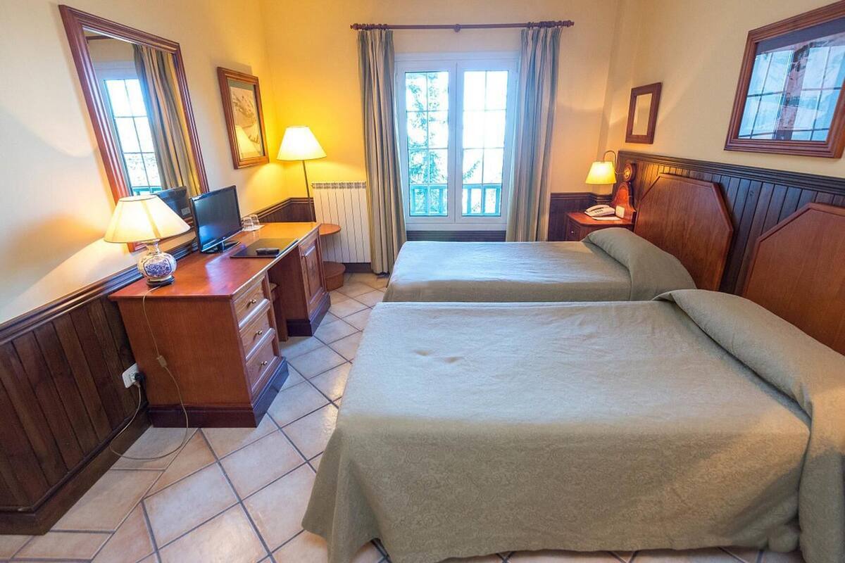 Una habitación de hotel con dos camas, un escritorio con computadora, una lámpara y un ventanal con cortinas.