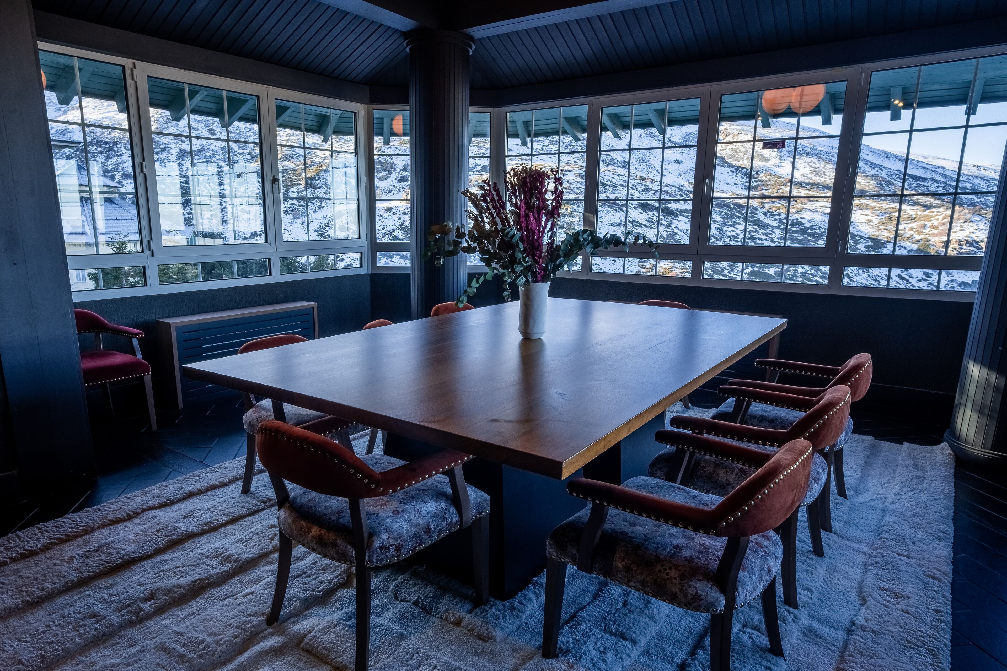 Une salle de conférence dotée d’une grande table et de six chaises, de grandes fenêtres offrant une vue sur une montagne enneigée, et d’un vase rempli de fleurs posé sur la table.