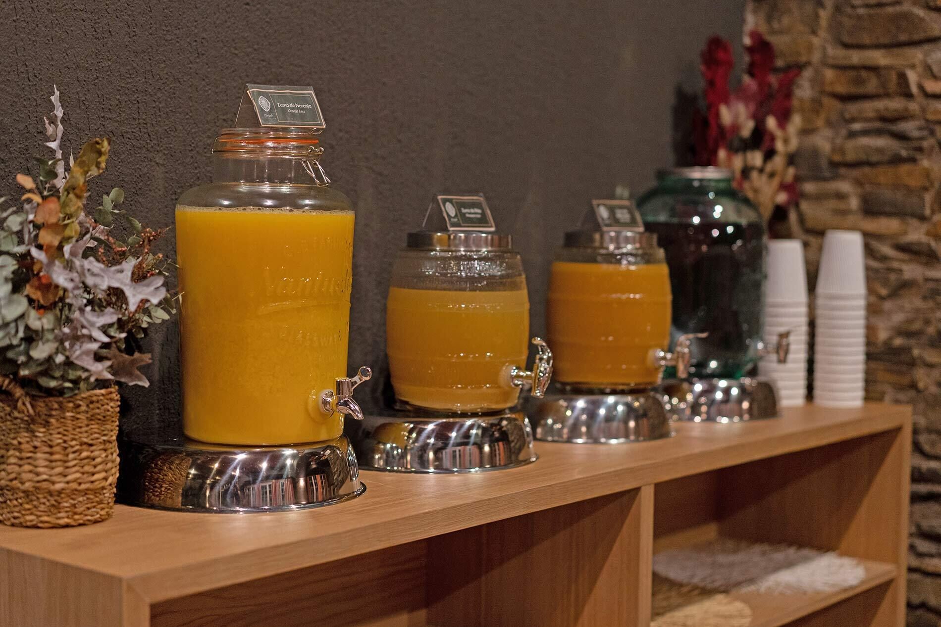 Una estación de bebidas con varios dispensadores de jugo de naranja, una cesta tejida y un florero con flores secas.