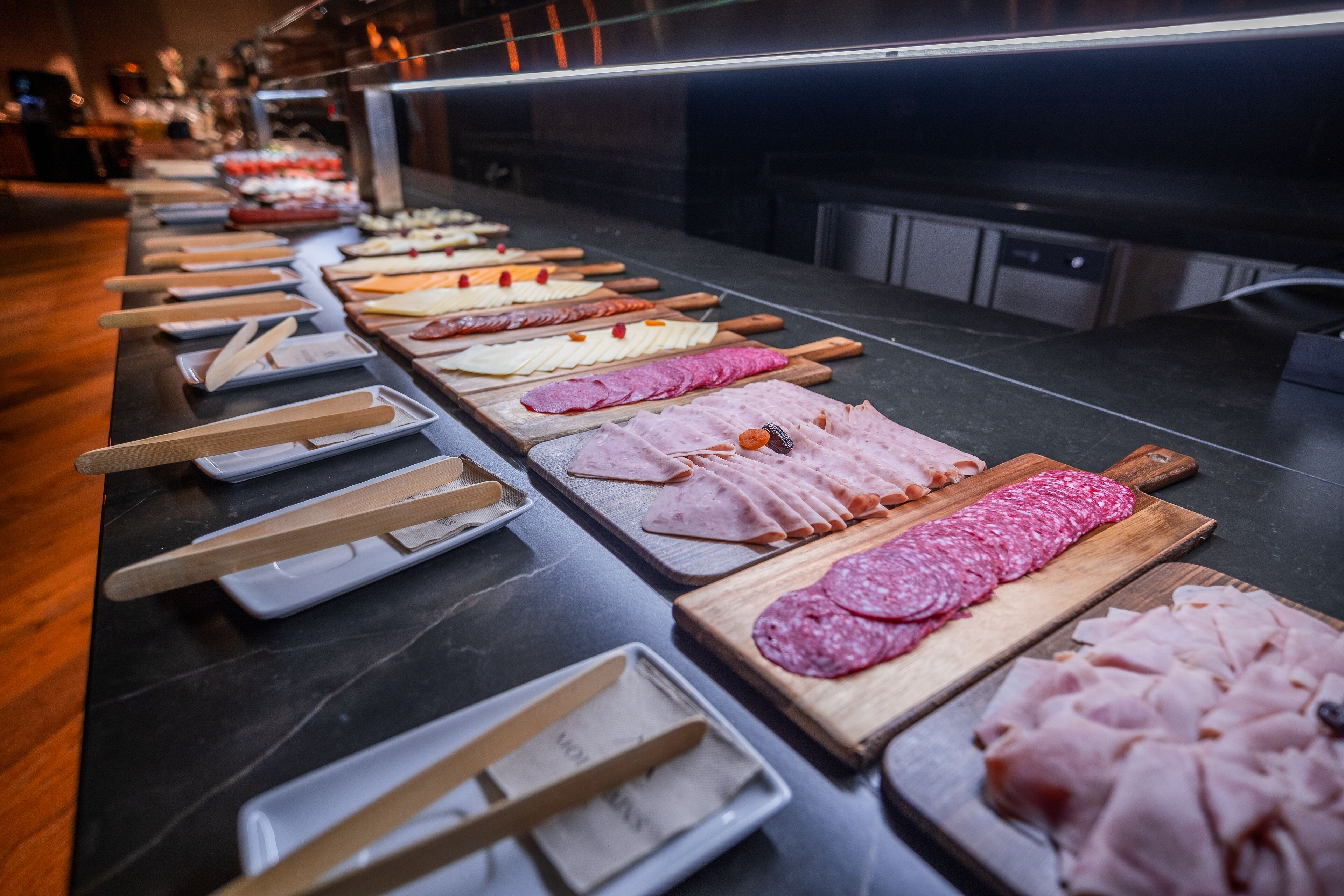 A buffet table with various types of deli meats and cheeses, wooden serving utensils, and white plates.